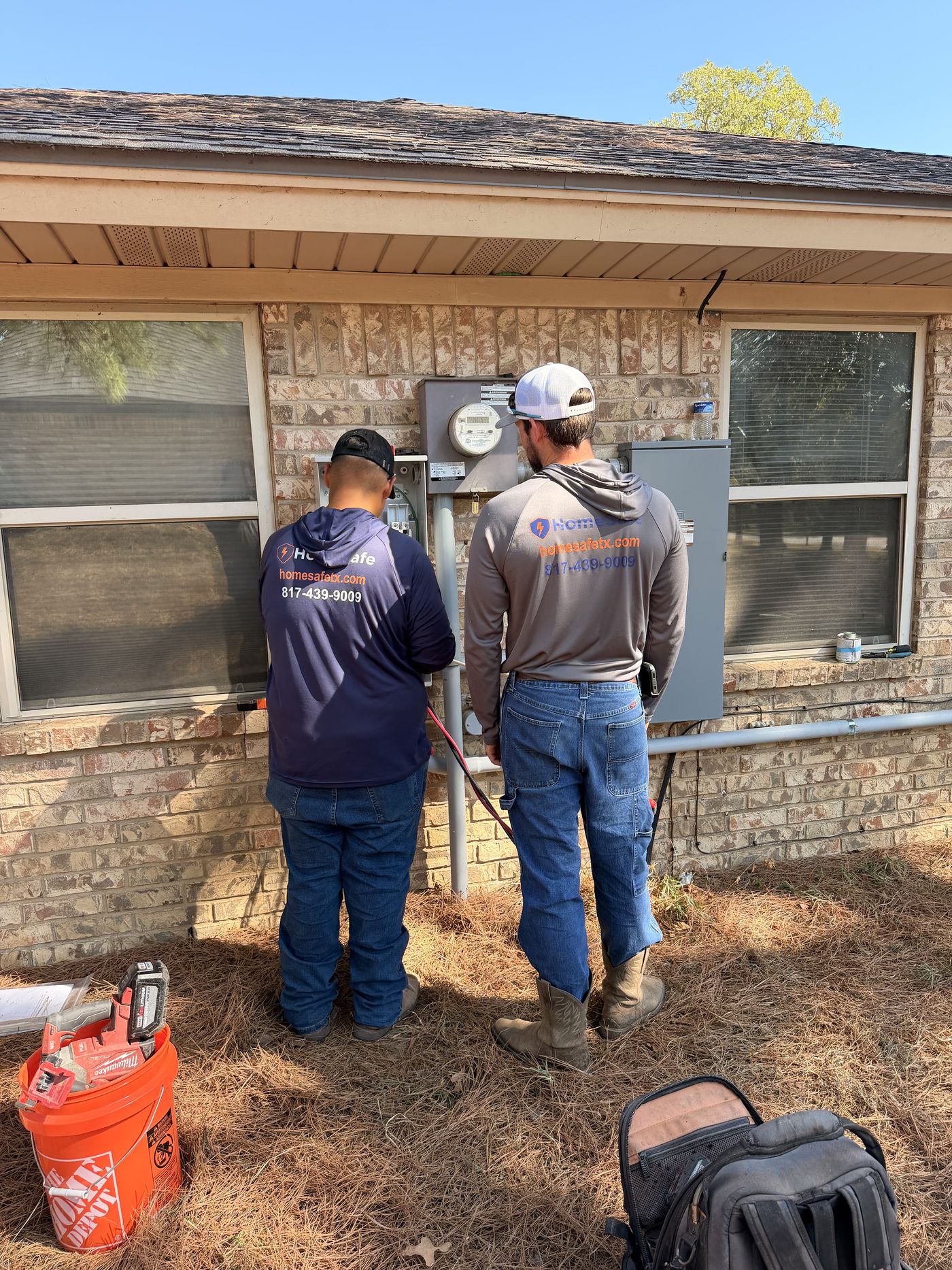 Two HomeSafe technicians working at the meter and electrical panel of an older brick home